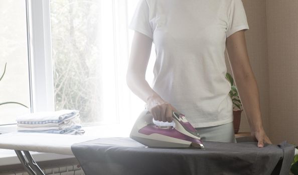 Smiling beautiful dark-skinned brunette woman in white t-shirt ironing blue skirt iron on an ironing board at home.