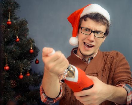 Man hipster in santa hat opens champagne on the background of Christmas tree