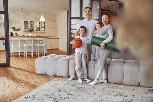 Full length portrait of joyful parents with son and daughter standing with sport equipment in living room