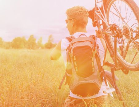 Cyclist man with backpack carries bicycle in green meadow in summer. Image with sunlight effect