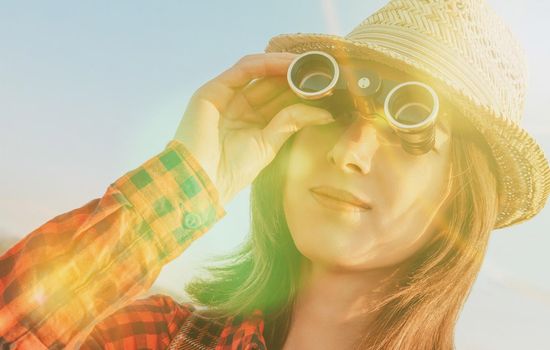 Smiling traveler young woman looking through binoculars outdoor. Image with sunlight effect