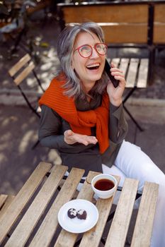 Emotional grey haired mature woman with glasses talks on mobile phone sitting at table on outdoors cafe terrace on autumn day