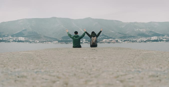 Happy traveler couple sitting on pier at bay in front of mountains and town. Space for text in lower part of image.