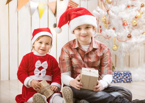 Kids in red santa hats sitting in decorated room with present boxes in hands