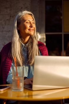 Cheerful senior Asian lady with works on contemporary laptop witting at small round table on outdoors cafe terrace at sunset