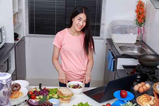 young woman mixing salad while cooking with laptop in kitchen 