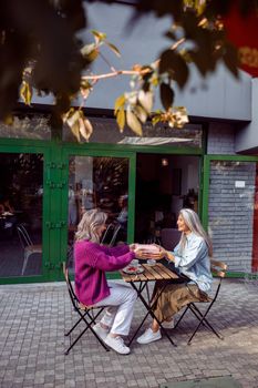 Positive senior women companions in elegant clothes hold gift box sitting table on outdoors cafe terrace on autumn day