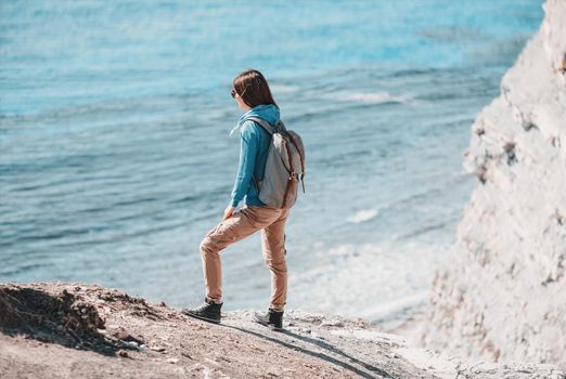 Young woman with backpack standing on coast and enjoying view of sea in summer