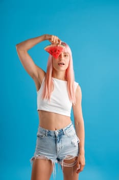 Cute young woman with pink hair holding a slice of watermelon over her eye, standing isolated over blue studio background. Beauty and health concept