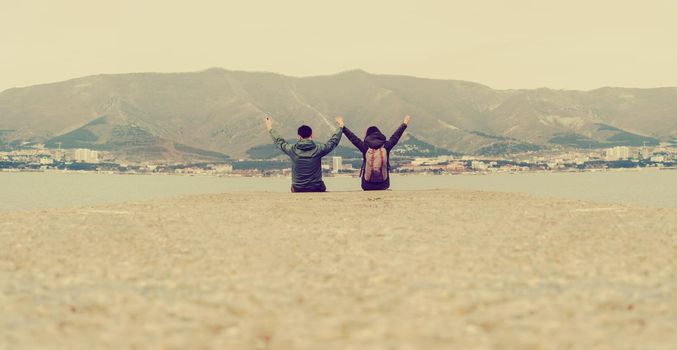 Happy loving traveler couple sitting on pier at bay in front of mountains and town. Space for text in lower part of image. Image with instagram color