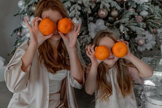 Mom and daughter hold tangerines at eye level instead of glasses. They are sitting under a decorated Christmas tree in their pajamas. The concept of Christmas, family holidays.
