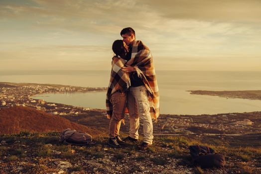 Loving couple wrapped in plaid standing on peak of mountain above bay at sunset