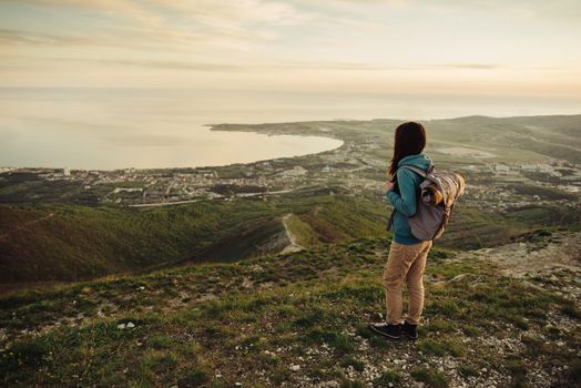 Traveler young woman with backpack standing on peak of mountain above the sea at sunset