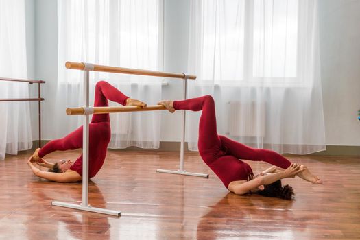 Two young womans fitness instructor in red Sportswear Leggings and Top stretching in the gym before pilates, on a yoga mat near the large window on a sunny day, female fitness yoga routine concept.