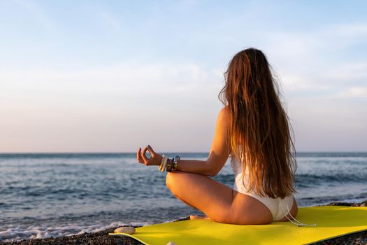 Young woman in swimsuit with long hair practicing stretching outdoors on yoga mat by the sea on a sunny day. Women's yoga fitness pilates routine. Healthy lifestyle, harmony and meditation concept.