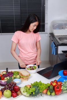 young woman mixing salad while cooking with laptop in kitchen 