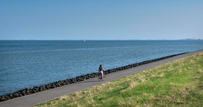 oudeschild, netherlands, 19 july 2021: woman rides bicycle on dike of wadden sea on dutch island of texel in summer under blue sky in holland