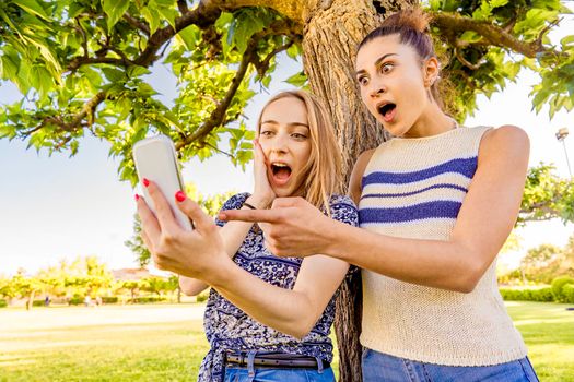 Two girls making surprised faces spreading mouth and eyes pointing and looking smartphone spending time in city park nature. Young women having fun with social network due to wi-fi mobile technology