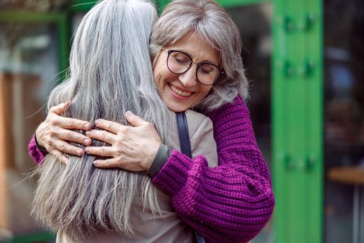 Happy senior woman with glasses in knitted jacket hugs best friend with long silver hair meeting on city street