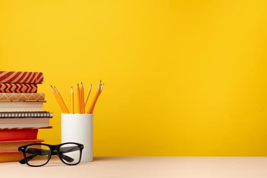 Cup of pencils and notepads on desk against yellow background, front view