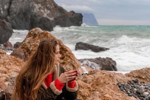 A young beautiful tourist girl is enjoying the autumn nature, sitting on a rock with a sea view and holding a cup of thermos in her hand. She is dressed in black jeans, boots, a red sweater and a jacket.