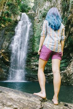 Explorer young woman standing on tree trunk and looking at waterfall in summer, rear view.