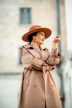 Outdoor fashion portrait of young elegant fashionable brunette woman, model in stylish hat, choker and light raincoat posing at sunset in European city