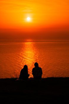 Two young woman in cute summer dress sitting on the stone, looking at golden sunset over the sea. Copy space. The concept of calmness, silence and unity with nature.