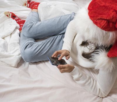 Beautiful happy brunette woman makes a selfie by phone in a white sweater and a Christmas hat and holds a Christmas present while lying in a white bed