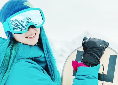 Smiling young woman with blue hair in protective sunglasses standing with snowboard, looking at camera. Snow mountains reflected in glasses.