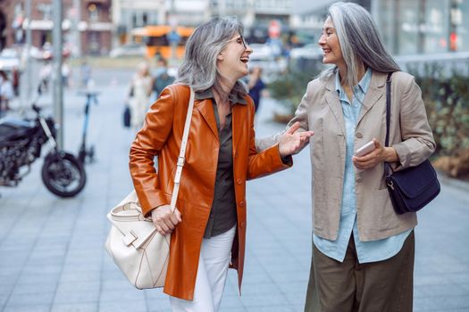 Positive senior women in stylish garment talk walking along modern city street on autumn day. Friends spend time together