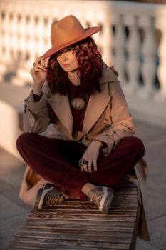 Outdoor fashion portrait of young elegant fashionable brunette woman, model in stylish hat, choker and light raincoat posing at sunset in European city