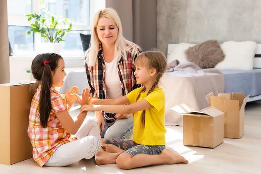 Caucasian family, woman and two girls sit on floor, unpack boxes and smile in new house. Behind them moving boxes.