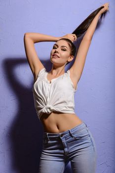 Sexy tanned brunette girl in a white vest and jeans raising his hands and gathered her hair in a ponytail on a blue background