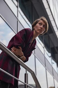 young woman with short hair in red checkered jacket outdoors in front of office building. portrait of caucasian female leaning on railings at street