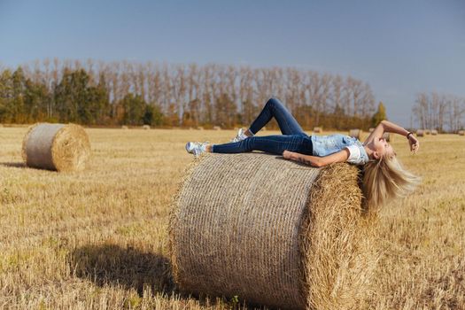 Beautiful girl villager posing in jeans on a bale of hay in a field