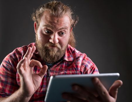 Man with beard and long hair wearing plaid shirt, holding tablet and showing ok sign, satisfied gesture, shoot in studio