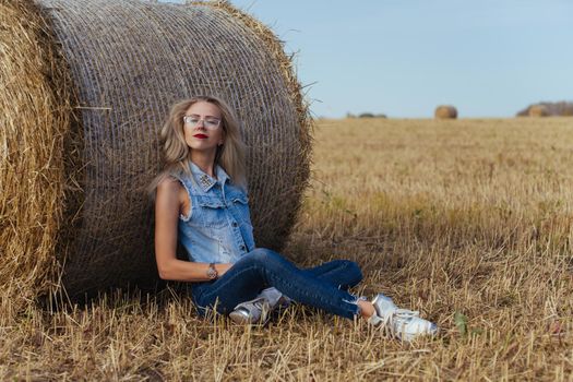 Beautiful girl villager posing in jeans near a bale of hay in a field