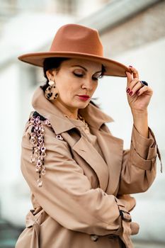 Outdoor fashion portrait of young elegant fashionable brunette woman, model in stylish hat, choker and light raincoat posing at sunset in European city