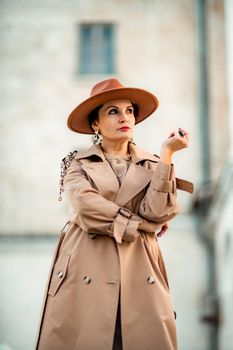 Outdoor fashion portrait of young elegant fashionable brunette woman, model in stylish hat, choker and light raincoat posing at sunset in European city