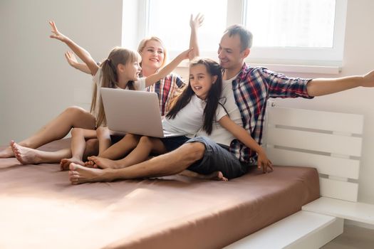Happy family concept. Beautiful mother and handsome father with their daughters spending time together at home and lying on bed with laptop.