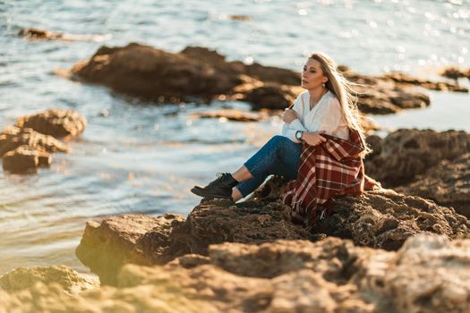 Attractive blonde Caucasian woman enjoying time on the beach at sunset, sitting in a blanket and looking to the side, with the sunset sky and sea in the background. Beach vacation