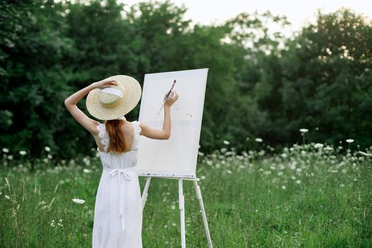 A woman in a white dress in a field with flowers paints a picture. High quality photo