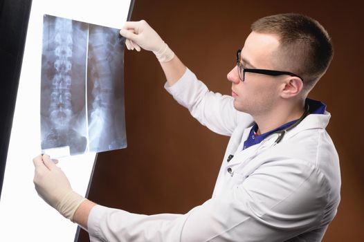 Young doctor radiologist in glasses and medical clothes looks at the lumen x-ray picture of the spine on a brown background