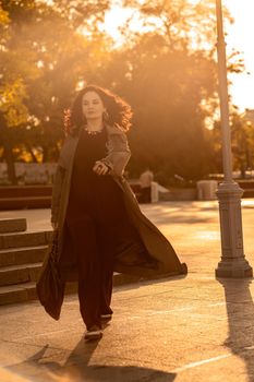 Outdoor fashion portrait of young elegant fashionable brunette woman, model in stylish hat, choker and light raincoat posing at sunset in European city