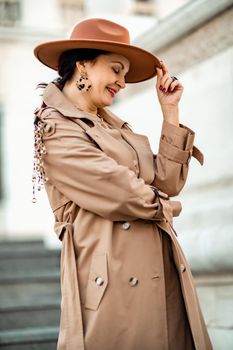 Outdoor fashion portrait of young elegant fashionable brunette woman, model in stylish hat, choker and light raincoat posing at sunset in European city