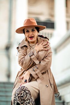 Outdoor fashion portrait of young elegant fashionable brunette woman, model in stylish hat, choker and light raincoat posing at sunset in European city