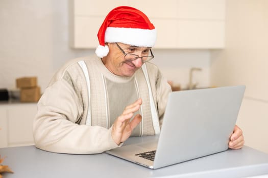 Elderly Gentleman Celebrating Christmas Online At Laptop Making Video Call With Distant Family Sitting In Kitchen At Home. New Year Holiday.