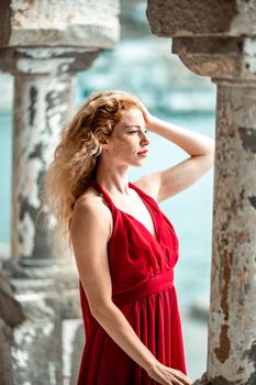 Outdoor portrait of a young beautiful natural redhead girl with freckles, long curly hair, in a red dress, posing against the background of the sea