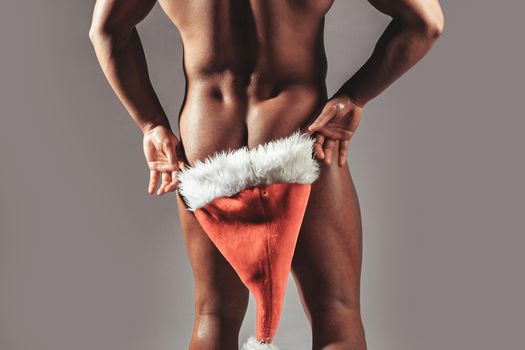 Sexy muscular gay man hiding buttocks butt, ass under red santa claus hat. Winter holidays celebration on grey.